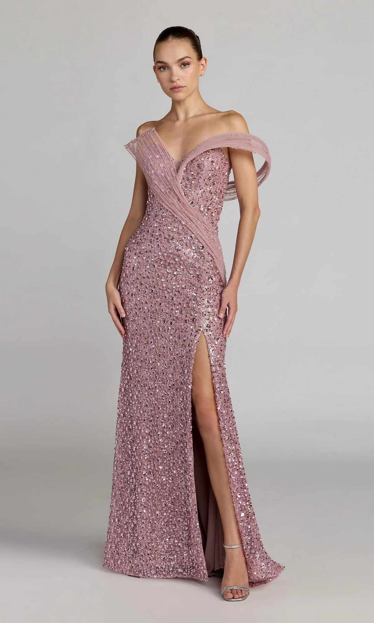 A woman wears the Mac Duggal 37085 Long Formal Dress, a floor-length pink sequin gown with an off-the-shoulder asymmetrical neckline and thigh-high slit, radiating couture-inspired elegance.